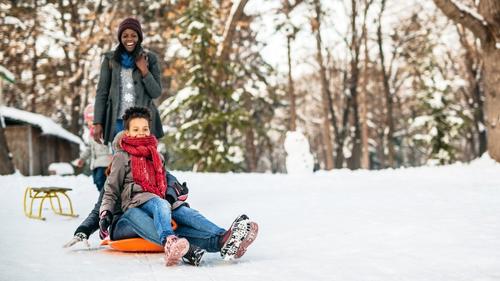 Two people sledding and one person standing on a winter trail.