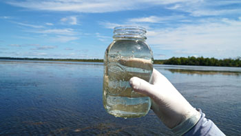 A white-gloved hand holds up a glass jar containing water with a lake in the background
