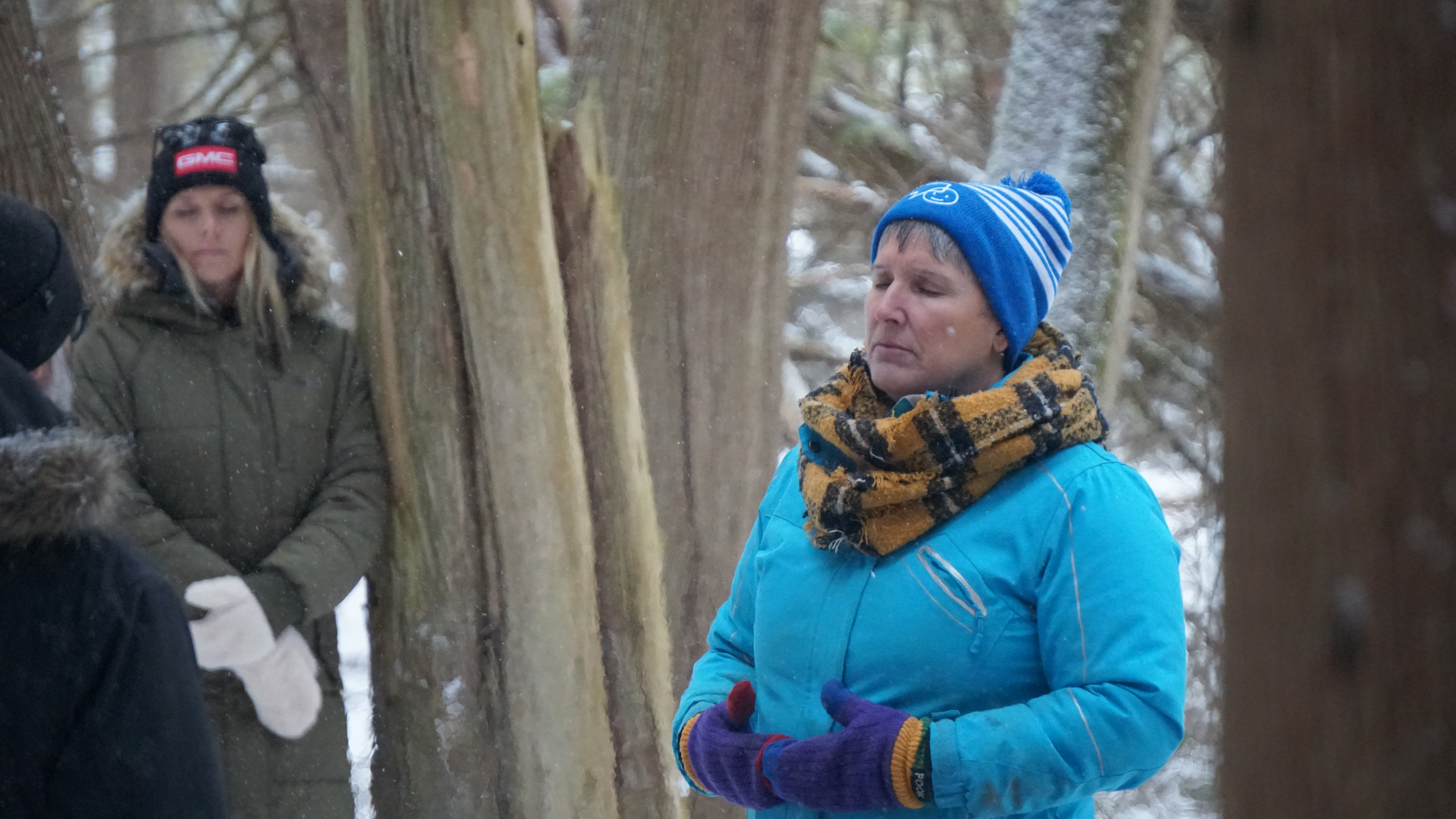 A person in a blue winter coat and blue hat stands with their eyes closed in the forest listening to the sounds of nature during a Forest Therapy experience.